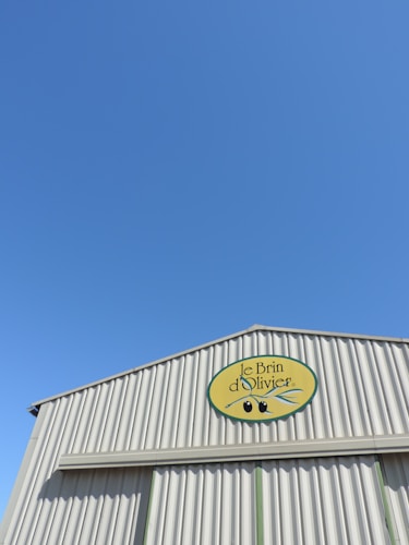 A building with corrugated metal siding and a logo displaying 'Le Brin d'Olivier' under a clear blue sky. The logo has an image of olives and an olive branch.
