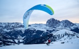 A person is paragliding on a snowy mountain landscape, with expansive views of distant peaks and snow-covered slopes. The sky is clear and the paraglider canopy is predominantly blue and green, contrasting with the white snow below. The scene depicts a sense of adventure and freedom amidst the vastness of nature.