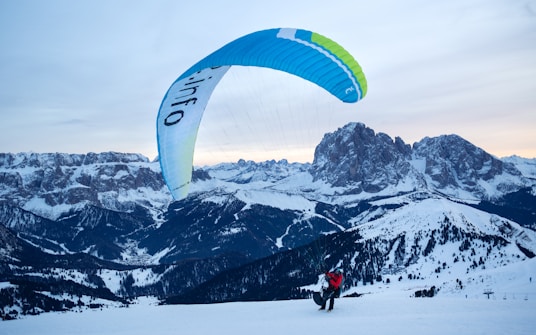 A person is paragliding on a snowy mountain landscape, with expansive views of distant peaks and snow-covered slopes. The sky is clear and the paraglider canopy is predominantly blue and green, contrasting with the white snow below. The scene depicts a sense of adventure and freedom amidst the vastness of nature.