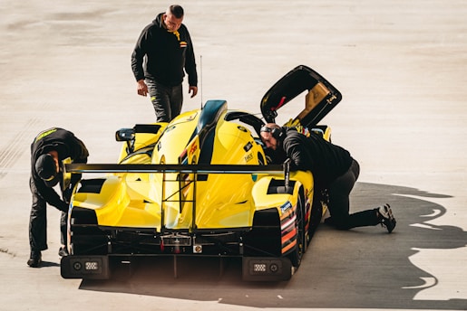 A group of people are inspecting and working on a bright yellow race car in a garage or pit area. One person is kneeling and looking closely at the car's interior, another is standing and supervising, while the third is bent over examining the rear tire area. The setting is well-lit and appears to be an outdoor or semi-outdoor space with a concrete floor.