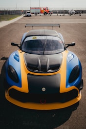 A brightly colored sports car with a predominantly yellow and blue exterior is parked on an asphalt surface in a racetrack environment. The car's hood is black with visible air vents, and a large spoiler is mounted at the rear. The windshield displays 'Lotus Cup Europe.' In the background, a fire truck and other vehicles are visible, along with a fenced area and a clear sky.