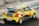 A yellow race car with black stripes speeding on a racetrack. The car is decorated with various sponsor logos and numbers, and there's a rear spoiler on its back. The setting appears to be a professional racing circuit.