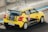 A yellow race car with black stripes speeding on a racetrack. The car is decorated with various sponsor logos and numbers, and there's a rear spoiler on its back. The setting appears to be a professional racing circuit.