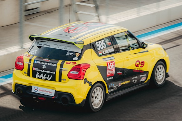 A yellow race car with black stripes speeding on a racetrack. The car is decorated with various sponsor logos and numbers, and there's a rear spoiler on its back. The setting appears to be a professional racing circuit.