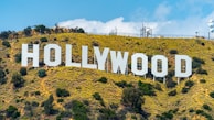 A large white sign spelling out 'HOLLYWOOD' set against a hillside covered with greenery and shrubs. The bright sky with scattered clouds forms the backdrop.