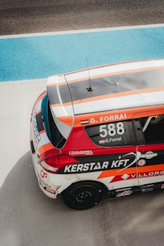 A race car featuring vibrant red, orange, and white colors is parked on a racing track. The car's roof displays the name 'G. Forrai' and the number '588', along with various sponsor logos and decals. The racing environment is highlighted by the blue and gray track surface surrounding the vehicle.
