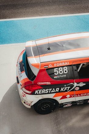 A race car featuring vibrant red, orange, and white colors is parked on a racing track. The car's roof displays the name 'G. Forrai' and the number '588', along with various sponsor logos and decals. The racing environment is highlighted by the blue and gray track surface surrounding the vehicle.