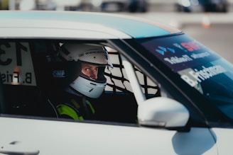 A person wearing a racing helmet sits inside a car, visible through the side window. The car is equipped with protective netting on the window and the driver's clothing is adorned with racing gear branding.
