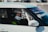 A person wearing a racing helmet sits inside a car, visible through the side window. The car is equipped with protective netting on the window and the driver's clothing is adorned with racing gear branding.