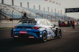A blue racing car is positioned on a paved track at a motorsport event. The car is adorned with various sponsor logos and a large rear spoiler. Spectators and event staff are visible in the background, along with a series of empty blue and white stands. Several flags are waving on poles, indicating an international setting.
