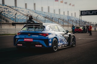 A blue racing car is positioned on a paved track at a motorsport event. The car is adorned with various sponsor logos and a large rear spoiler. Spectators and event staff are visible in the background, along with a series of empty blue and white stands. Several flags are waving on poles, indicating an international setting.