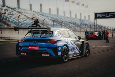A blue racing car is positioned on a paved track at a motorsport event. The car is adorned with various sponsor logos and a large rear spoiler. Spectators and event staff are visible in the background, along with a series of empty blue and white stands. Several flags are waving on poles, indicating an international setting.