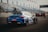A blue racing car is positioned on a paved track at a motorsport event. The car is adorned with various sponsor logos and a large rear spoiler. Spectators and event staff are visible in the background, along with a series of empty blue and white stands. Several flags are waving on poles, indicating an international setting.