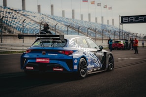 A blue racing car is positioned on a paved track at a motorsport event. The car is adorned with various sponsor logos and a large rear spoiler. Spectators and event staff are visible in the background, along with a series of empty blue and white stands. Several flags are waving on poles, indicating an international setting.