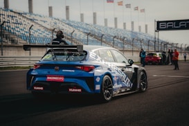 A blue racing car is positioned on a paved track at a motorsport event. The car is adorned with various sponsor logos and a large rear spoiler. Spectators and event staff are visible in the background, along with a series of empty blue and white stands. Several flags are waving on poles, indicating an international setting.