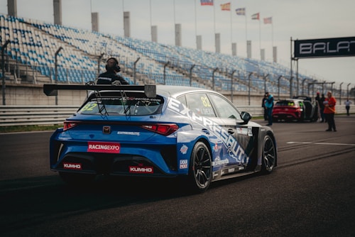 A blue racing car is positioned on a paved track at a motorsport event. The car is adorned with various sponsor logos and a large rear spoiler. Spectators and event staff are visible in the background, along with a series of empty blue and white stands. Several flags are waving on poles, indicating an international setting.