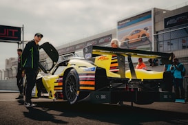 A bright yellow race car with black and red details is parked on a racing circuit. Several people are gathered around the car, some wearing caps and jackets, suggesting involvement with racing or as team members. The background includes fenced tracks and large advertisements featuring other vehicles.