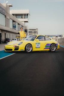 A yellow sports car with colorful stripes and exposed sponsor logos is parked on a race track. A person wearing a matching yellow racing suit stands beside the vehicle, holding a helmet. The background shows a modern building with large windows, typical of a raceway setting.