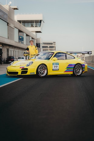 A yellow sports car with colorful stripes and exposed sponsor logos is parked on a race track. A person wearing a matching yellow racing suit stands beside the vehicle, holding a helmet. The background shows a modern building with large windows, typical of a raceway setting.
