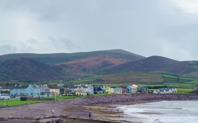 A coastal scene features a row of colorful houses along the shoreline with grassy areas in front. In the background, rolling hills with patches of green and brown rise into a cloudy sky. The foreground includes a rocky beach with waves gently lapping at the shore.