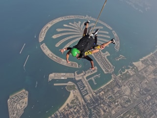 Aerial view of a person skydiving above a palm-shaped artificial archipelago surrounded by the ocean, with a coastal cityscape visible below.