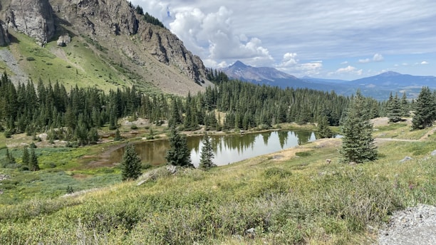 A serene mountain landscape features a reflective pond surrounded by lush greenery and sparse pine trees. A rocky cliff rises to the left, while distant mountains are visible under a partly cloudy sky.
