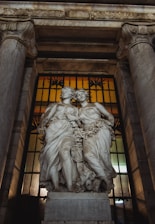 Two intricately carved marble statues are depicted, portraying figures adorned with floral garlands. These statues are positioned in an architectural setting with tall columns on each side and a decorative iron gate with amber-tinted glass behind them.