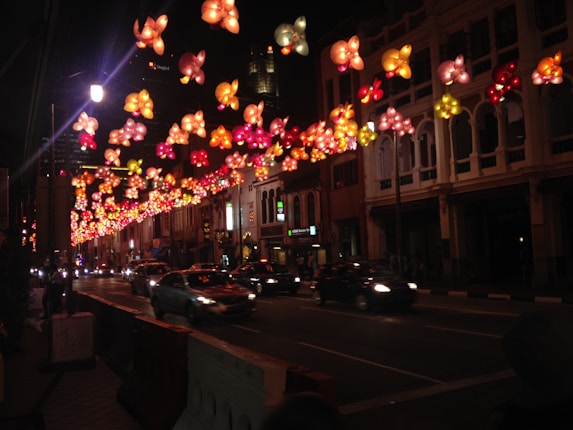 A nighttime city street decorated with colorful illuminated flower-shaped lanterns hanging above. The street is lined with multi-story buildings and several cars are driving by. The scene is lively and festive with vivid lighting against the dark sky.