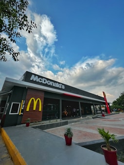 A McDonald's restaurant with a modern design, featuring an outdoor seating area. The large McDonald's sign is prominently displayed above. There are a few potted plants along the sidewalk, and the sky is partly cloudy with patches of blue.