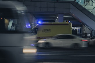 A nighttime urban scene featuring a yellow ambulance with emergency lights on, parked near a building. Cars are driving past swiftly, creating a motion blur effect. The environment appears to be an urban city street under artificial lighting.