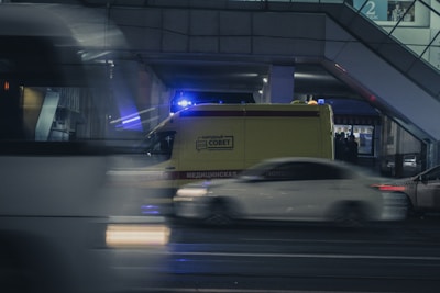 A nighttime urban scene featuring a yellow ambulance with emergency lights on, parked near a building. Cars are driving past swiftly, creating a motion blur effect. The environment appears to be an urban city street under artificial lighting.