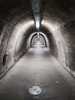 A long, narrow underground tunnel with curved walls and a concrete floor. The tunnel is illuminated by a series of lights attached to the ceiling, running the entire length of the tunnel. The walls appear to be old and weathered, with various markings and some minor graffiti. There is a circular manhole cover on the floor near the foreground.