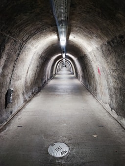 A long, narrow underground tunnel with curved walls and a concrete floor. The tunnel is illuminated by a series of lights attached to the ceiling, running the entire length of the tunnel. The walls appear to be old and weathered, with various markings and some minor graffiti. There is a circular manhole cover on the floor near the foreground.