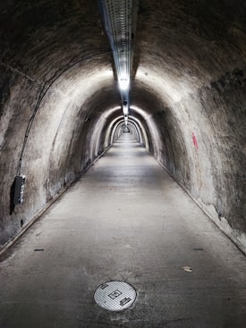 A long, narrow underground tunnel with curved walls and a concrete floor. The tunnel is illuminated by a series of lights attached to the ceiling, running the entire length of the tunnel. The walls appear to be old and weathered, with various markings and some minor graffiti. There is a circular manhole cover on the floor near the foreground.
