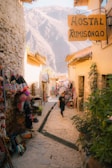 A narrow cobblestone alley lined with craft stalls displaying vibrant textiles and handcrafted items. The alleyway leads toward a backdrop of large, majestic mountains. A person walks down the path carrying a shopping bag, while sunlight casts warm tones over the rustic buildings.