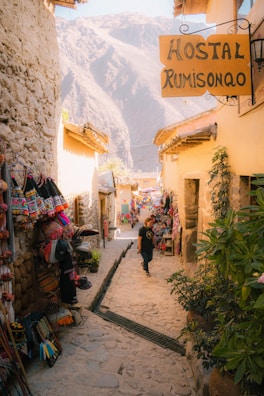 A narrow cobblestone alley lined with craft stalls displaying vibrant textiles and handcrafted items. The alleyway leads toward a backdrop of large, majestic mountains. A person walks down the path carrying a shopping bag, while sunlight casts warm tones over the rustic buildings.