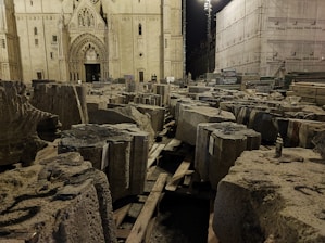 A Gothic-style cathedral facade is lit at night, with large stone blocks stacked and scattered on wooden pallets in the foreground. Scaffolding and construction materials are seen to the right, indicating renovation or restoration work is underway.