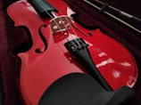 A red violin is resting in a dark velvet-lined case. The strings and fine-tuners are visible, highlighting the instrument's glossy finish. The bow is placed beside the violin.