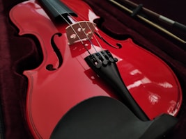 A red violin is resting in a dark velvet-lined case. The strings and fine-tuners are visible, highlighting the instrument's glossy finish. The bow is placed beside the violin.