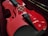 A red violin is resting in a dark velvet-lined case. The strings and fine-tuners are visible, highlighting the instrument's glossy finish. The bow is placed beside the violin.