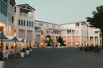 A modern, elegant building complex with white facades, large windows, and balconies. The architecture features striped awnings in various pastel colors and is surrounded by palm trees and outdoor seating areas. People can be seen relaxing in the open courtyard under the early evening sky, contributing to a serene atmosphere.