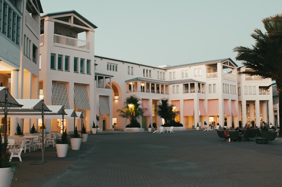 A modern, elegant building complex with white facades, large windows, and balconies. The architecture features striped awnings in various pastel colors and is surrounded by palm trees and outdoor seating areas. People can be seen relaxing in the open courtyard under the early evening sky, contributing to a serene atmosphere.