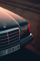 A classic Mercedes-Benz car with its front end prominently displayed, featuring the iconic star emblem on the hood. The image captures the detail of the grille and headlights, bathed in warm, diffused sunlight casting long shadows across the ground.