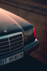 A classic Mercedes-Benz car with its front end prominently displayed, featuring the iconic star emblem on the hood. The image captures the detail of the grille and headlights, bathed in warm, diffused sunlight casting long shadows across the ground.