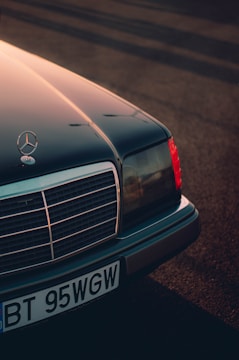 A classic Mercedes-Benz car with its front end prominently displayed, featuring the iconic star emblem on the hood. The image captures the detail of the grille and headlights, bathed in warm, diffused sunlight casting long shadows across the ground.