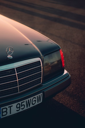 A classic Mercedes-Benz car with its front end prominently displayed, featuring the iconic star emblem on the hood. The image captures the detail of the grille and headlights, bathed in warm, diffused sunlight casting long shadows across the ground.
