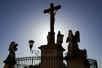 A silhouette of a large cross is centered against a bright sky, flanked by statues of angels. The scene is enclosed by ornate fencing and lit by traditional street lamps. A small bird is visible in the top of the frame, adding depth to the serene atmosphere.