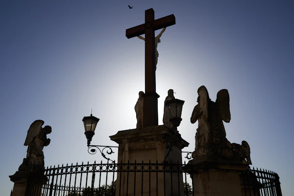 A silhouette of a large cross is centered against a bright sky, flanked by statues of angels. The scene is enclosed by ornate fencing and lit by traditional street lamps. A small bird is visible in the top of the frame, adding depth to the serene atmosphere.