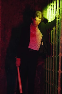 A person wearing a suit leans against a metal gate, holding a wooden bat. Dramatic lighting casts red and green tones across the scene, creating a tense atmosphere. The person's white shirt is stained, contributing to the overall moody ambiance.