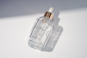 An elegant bottle of serum with droplets reflecting sunlight on a wooden surface with herbs.
